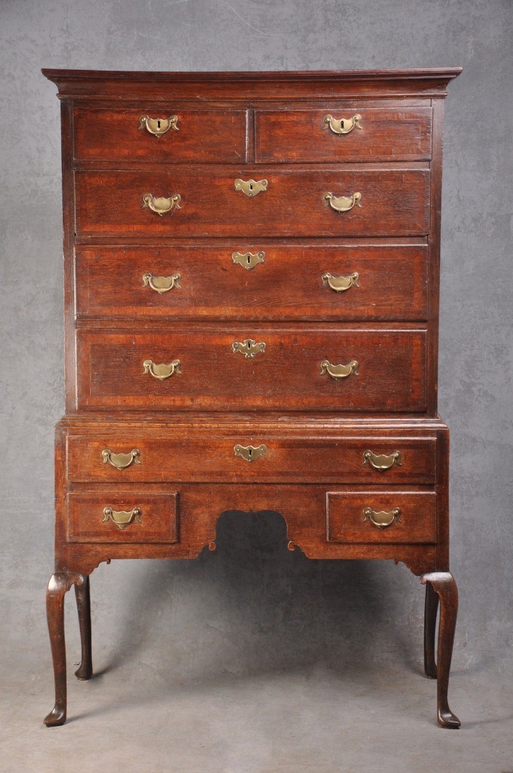 An early 18th-century oak chest on stand featuring two-part construction, multiple drawers with brass drop handles, a shaped apron, and slender cabriole legs.