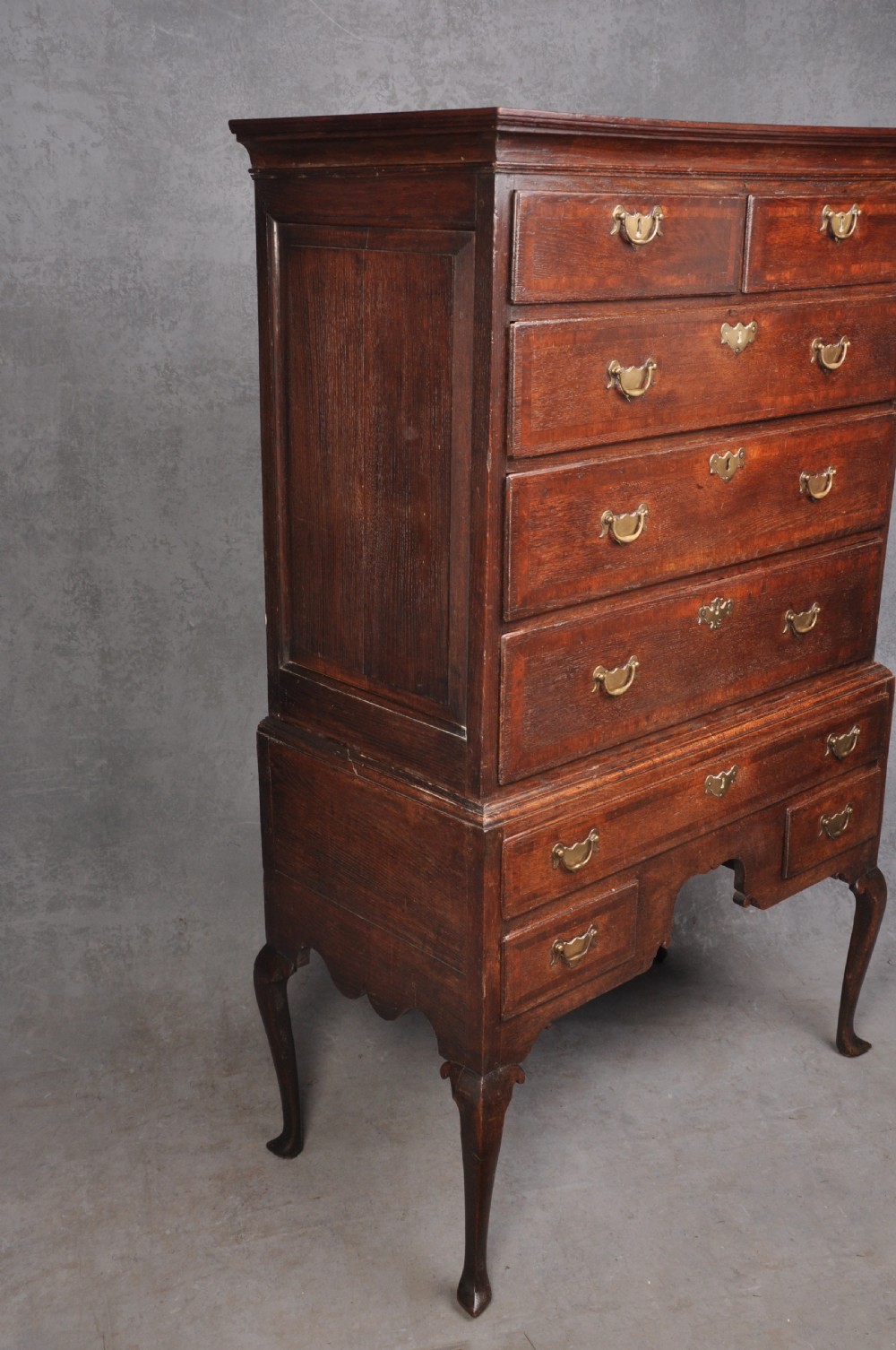 An early 18th-century oak chest on stand featuring two-part construction, multiple drawers with brass drop handles, a shaped apron, and slender cabriole legs. View X