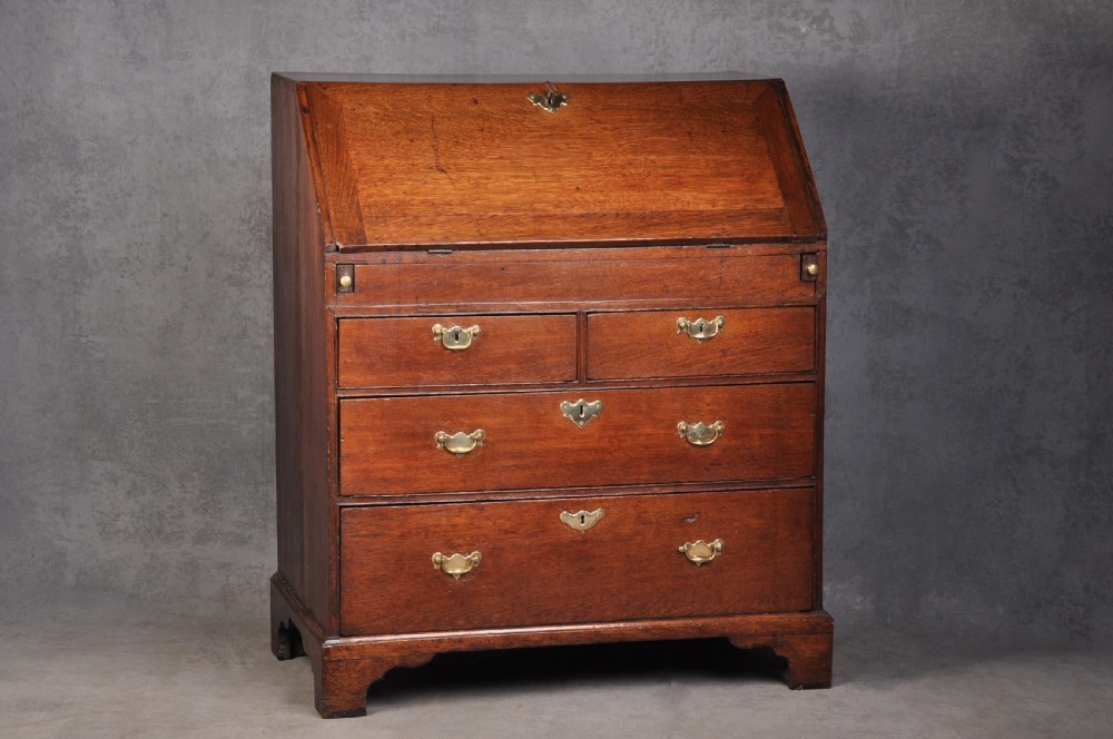 An early eighteenth-century oak bureau featuring a slanted drop-front desk above graduated drawers with brass drop handles and bracket feet.