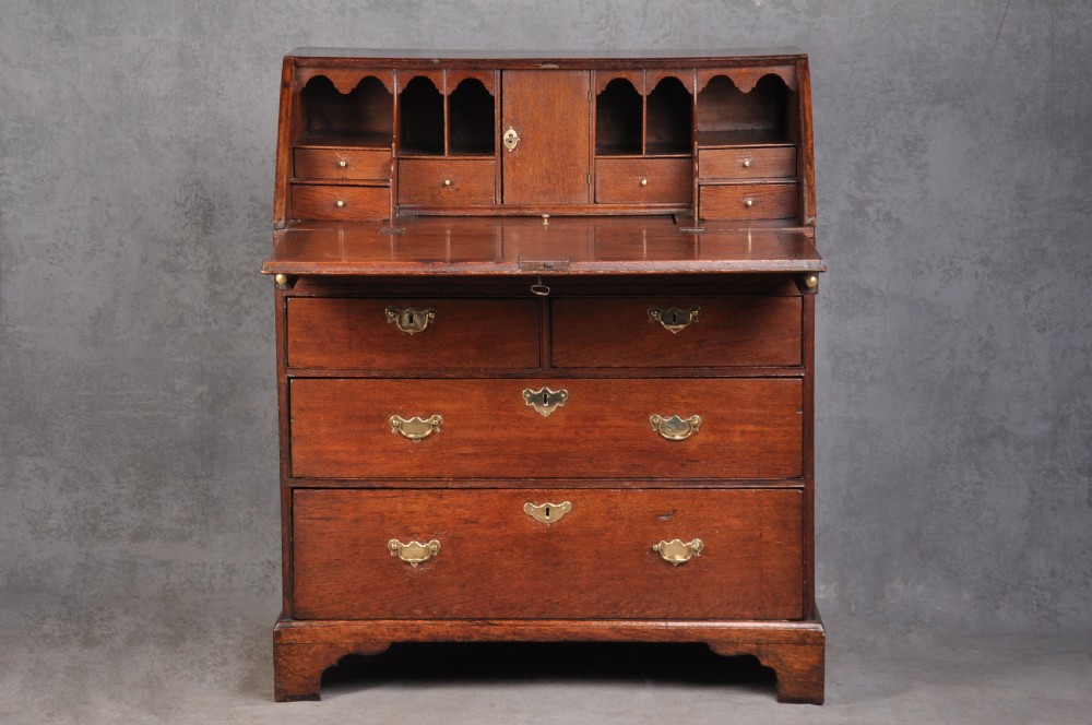 An early eighteenth-century oak bureau featuring a slanted drop-front desk above graduated drawers with brass drop handles and bracket feet. View II