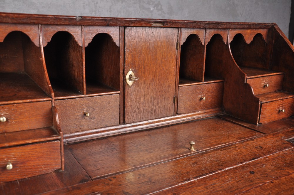 An early eighteenth-century oak bureau featuring a slanted drop-front desk above graduated drawers with brass drop handles and bracket feet. View IV