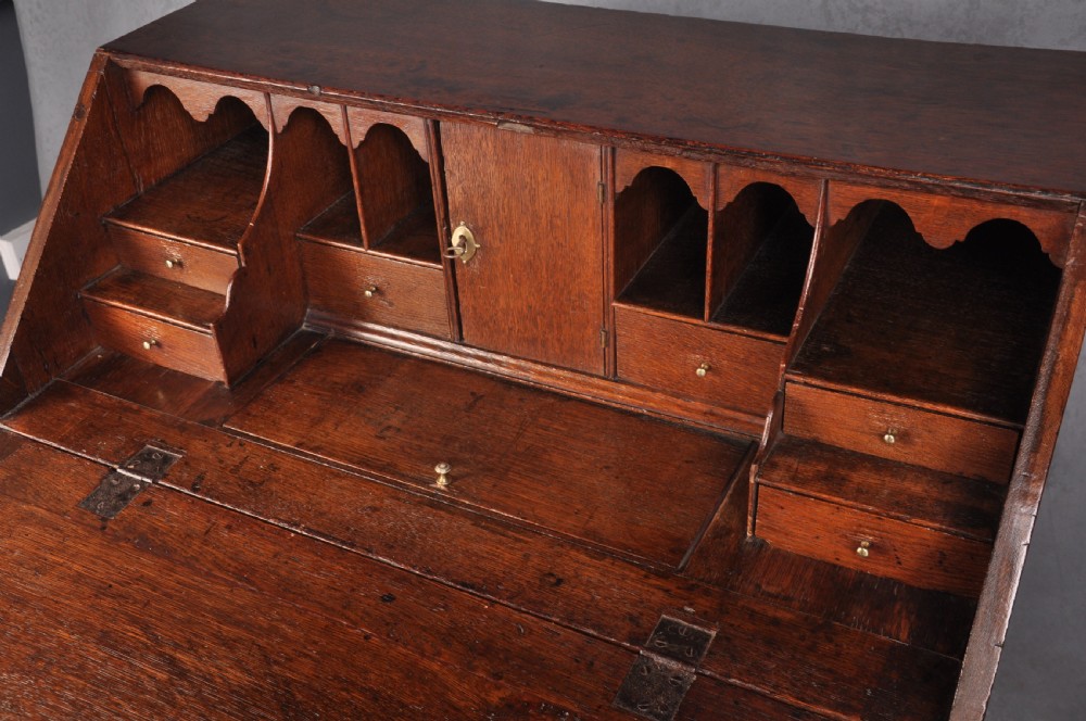 An early eighteenth-century oak bureau featuring a slanted drop-front desk above graduated drawers with brass drop handles and bracket feet. View V