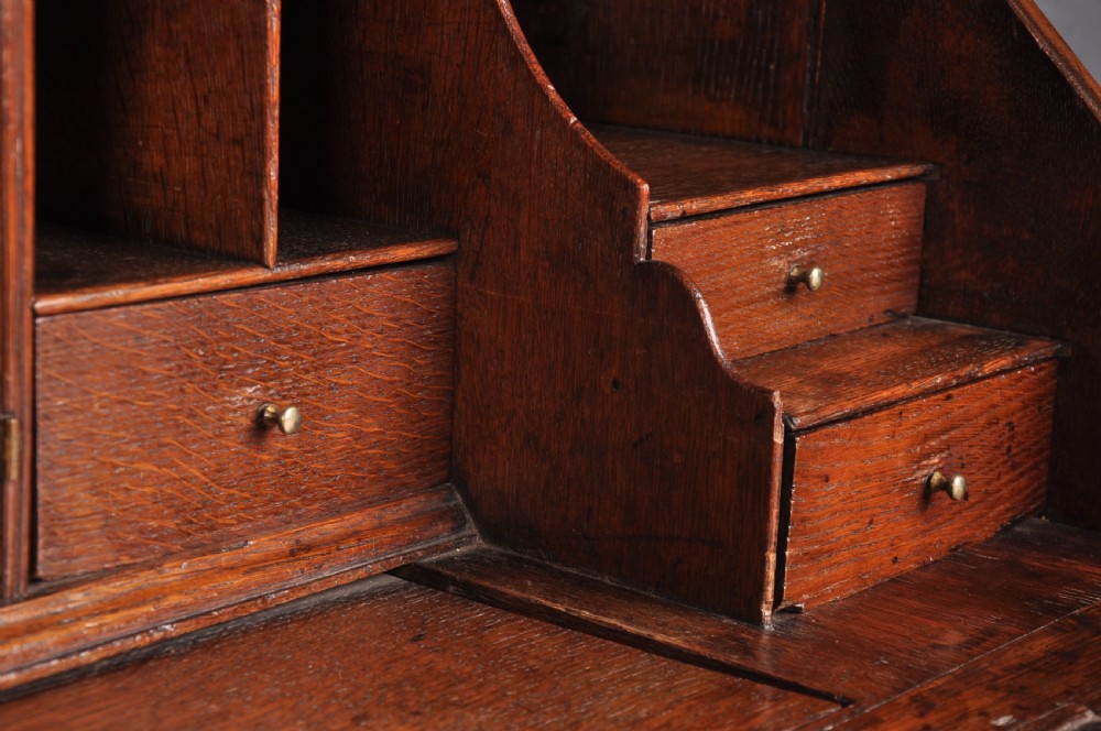 An early eighteenth-century oak bureau featuring a slanted drop-front desk above graduated drawers with brass drop handles and bracket feet. View VI