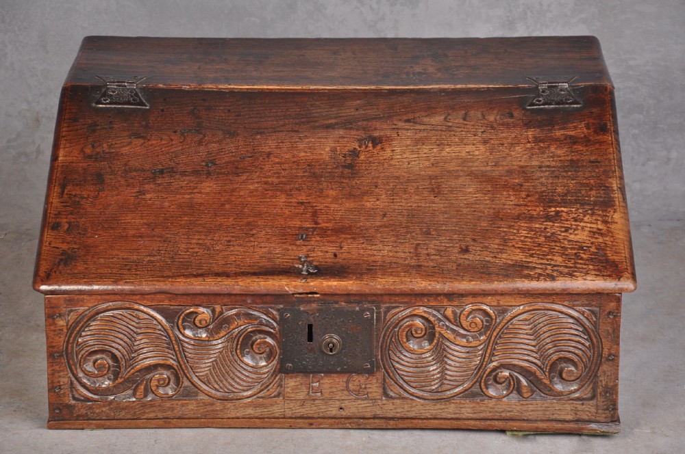 A 17th-century carved oak desk box featuring a sloped writing lid, iron hinges, iron lock plate, and hand-carved scroll motifs across the front panel.