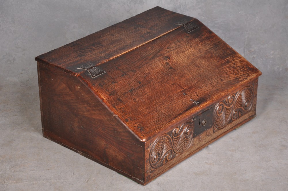 A 17th-century carved oak desk box featuring a sloped writing lid, iron hinges, iron lock plate, and hand-carved scroll motifs across the front panel. View V