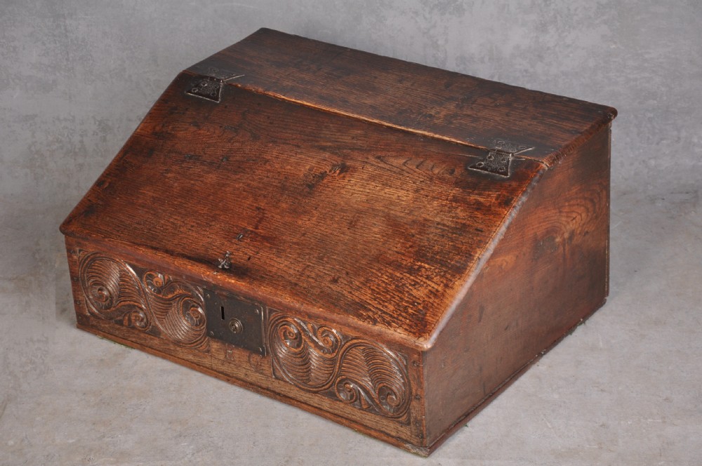 A 17th-century carved oak desk box featuring a sloped writing lid, iron hinges, iron lock plate, and hand-carved scroll motifs across the front panel. View VI