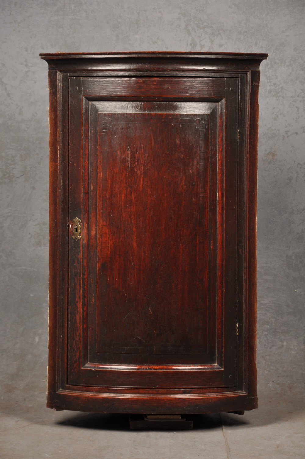 An early eighteenth-century dark oak bow-fronted corner cupboard featuring a curved fielded panel door, brass escutcheon, and moulded cornice.