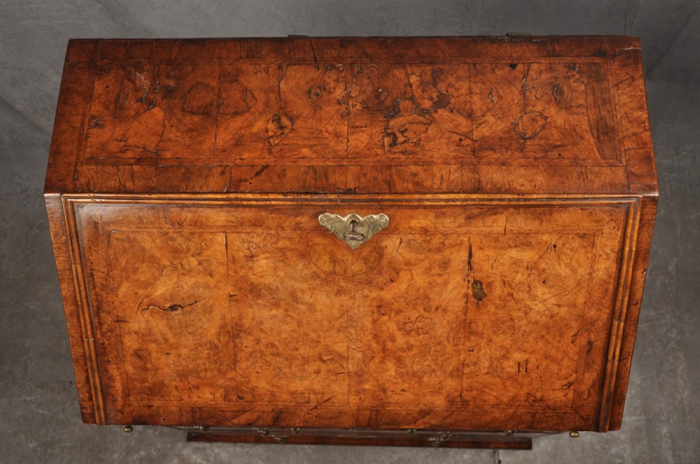 An early eighteenth-century burr walnut bureau featuring a sloped fall-front writing flap above four graduated drawers with brass drop handles and bracket feet. View XI