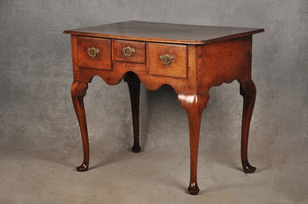 An early 18th-century red walnut lowboy table featuring three drawers with brass handles, a scalloped apron, and four cabriole legs on pad feet.