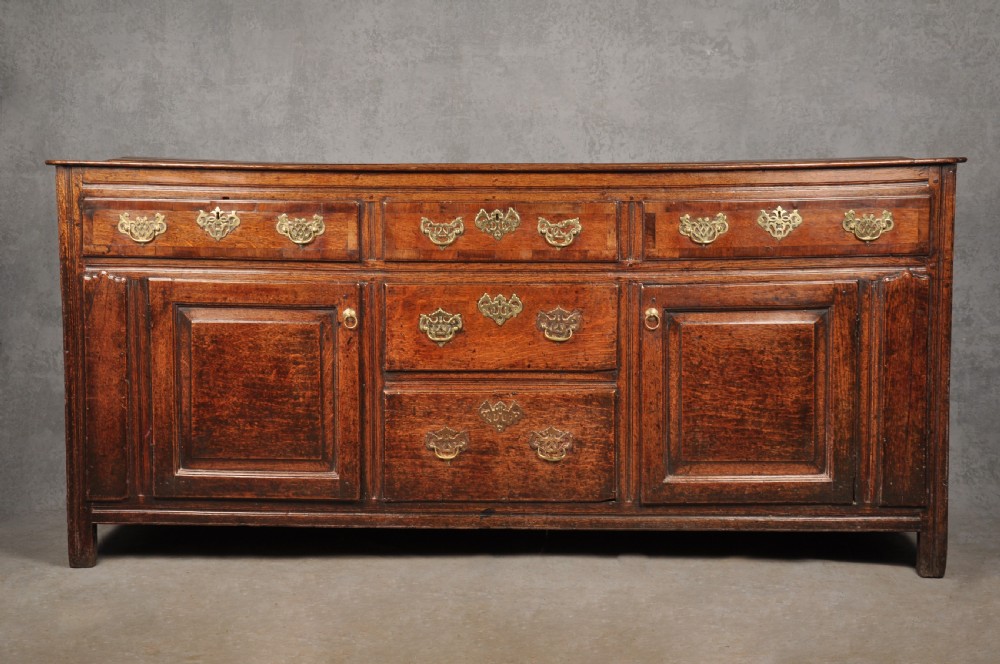 An early 18th-century dark oak dresser base featuring fielded panel cupboard doors, multiple drawers, and brass backplate handles.