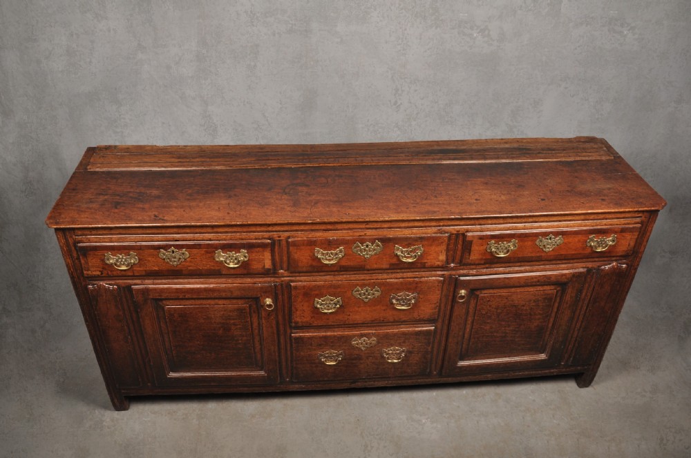An early 18th-century dark oak dresser base featuring fielded panel cupboard doors, multiple drawers, and brass backplate handles. View II