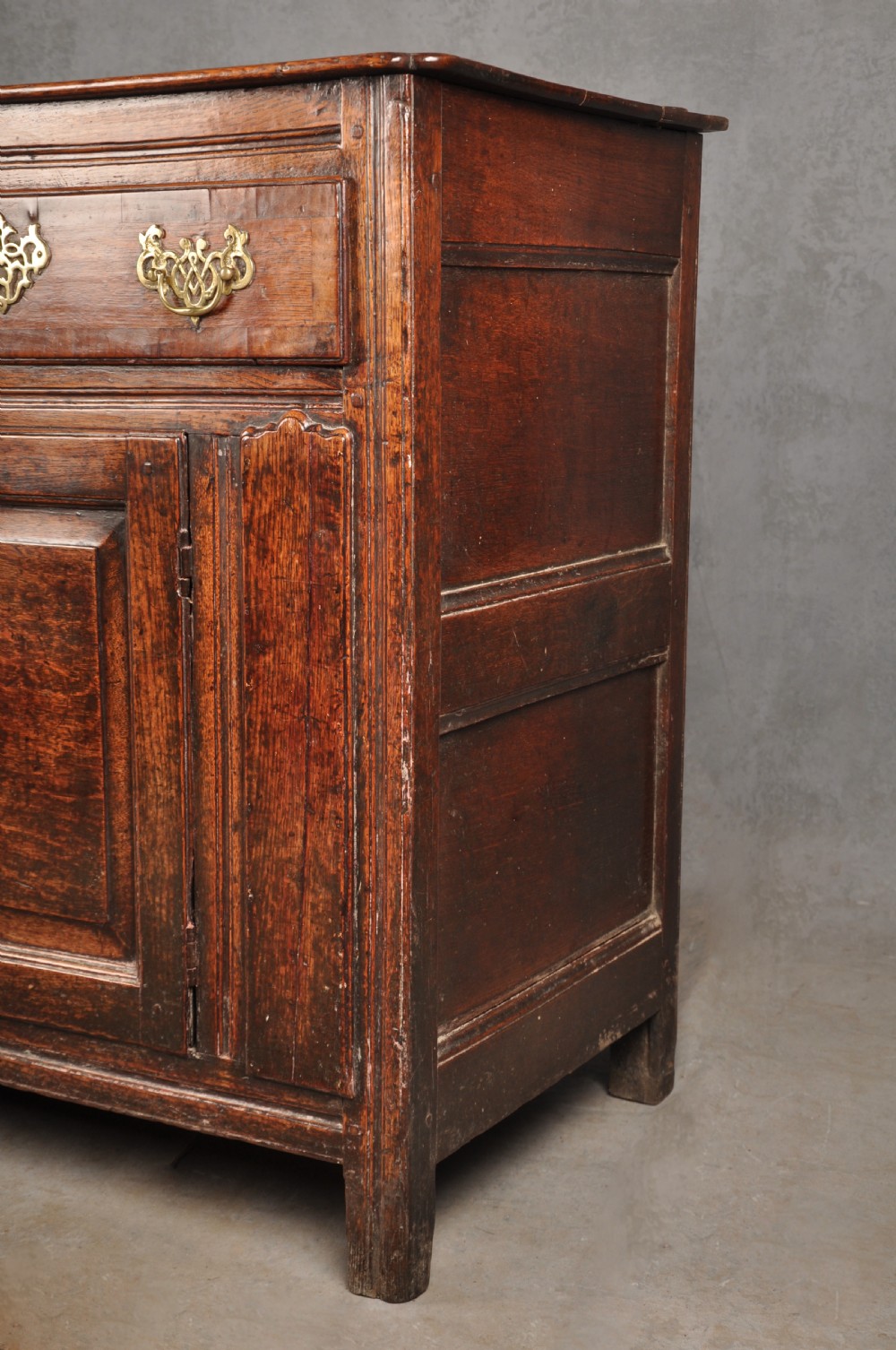 An early 18th-century dark oak dresser base featuring fielded panel cupboard doors, multiple drawers, and brass backplate handles. View III