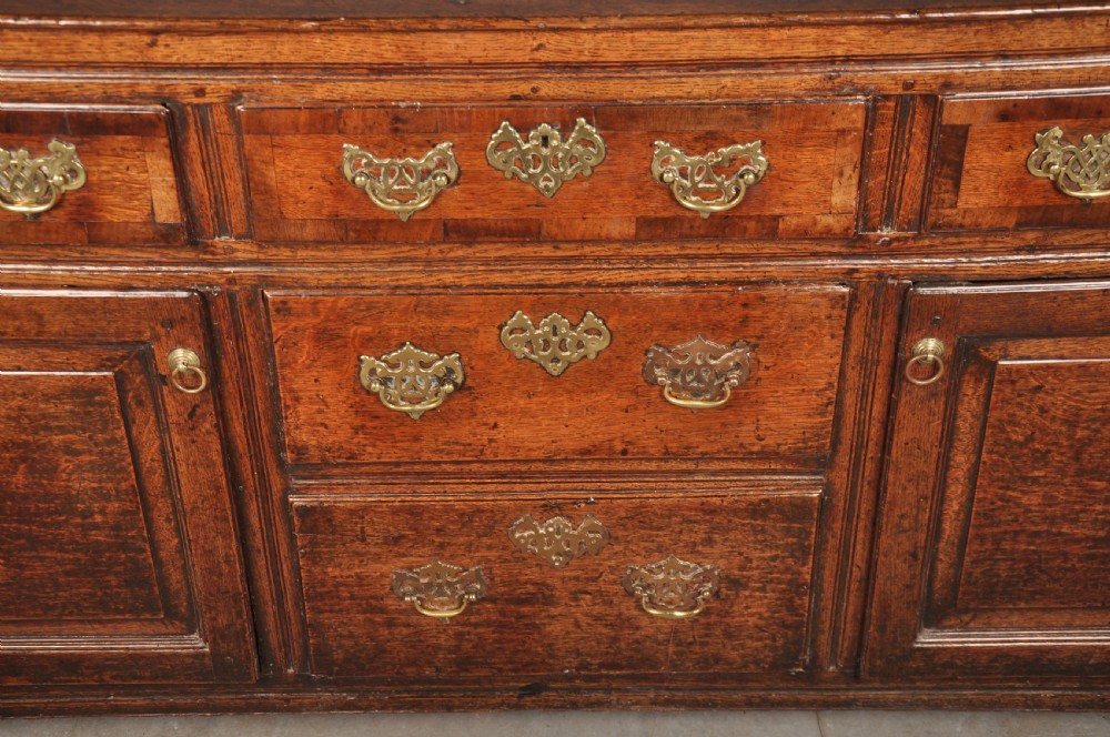 An early 18th-century dark oak dresser base featuring fielded panel cupboard doors, multiple drawers, and brass backplate handles. View V
