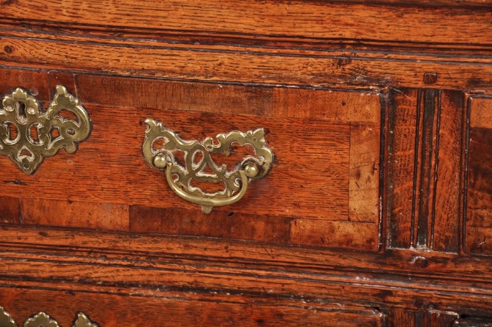 An early 18th-century dark oak dresser base featuring fielded panel cupboard doors, multiple drawers, and brass backplate handles. View VI