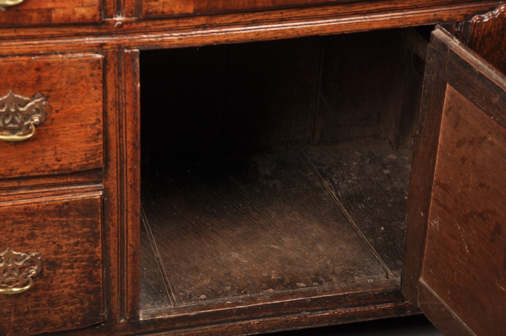 An early 18th-century dark oak dresser base featuring fielded panel cupboard doors, multiple drawers, and brass backplate handles. View XI