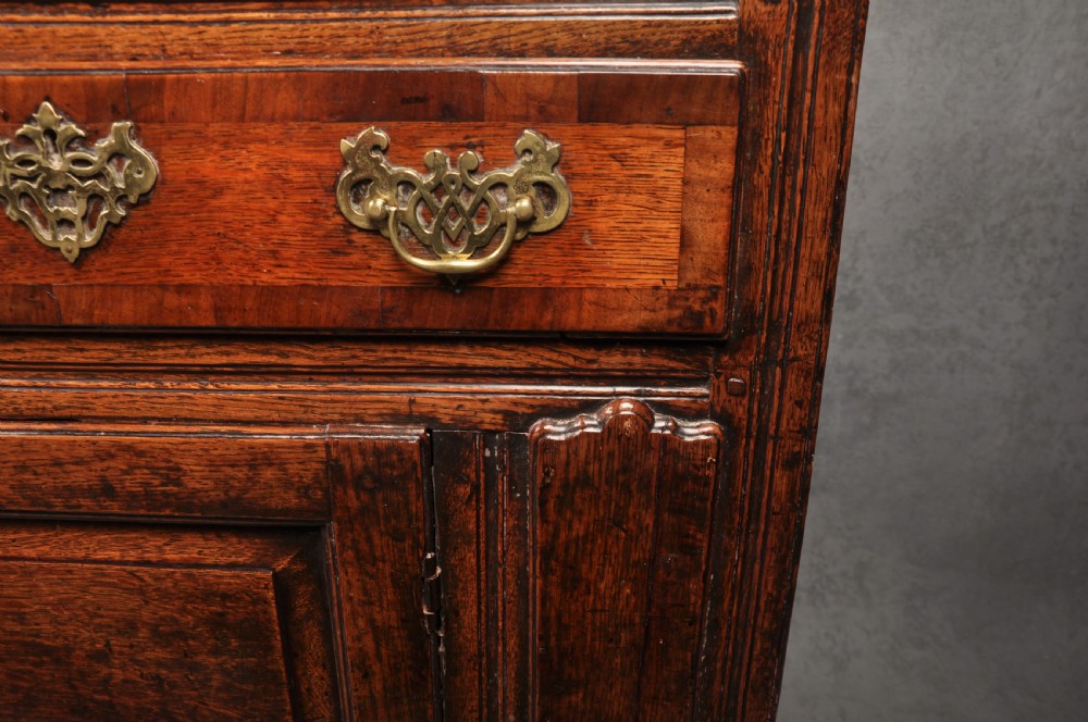 An early 18th-century dark oak dresser base featuring fielded panel cupboard doors, multiple drawers, and brass backplate handles. View XIII