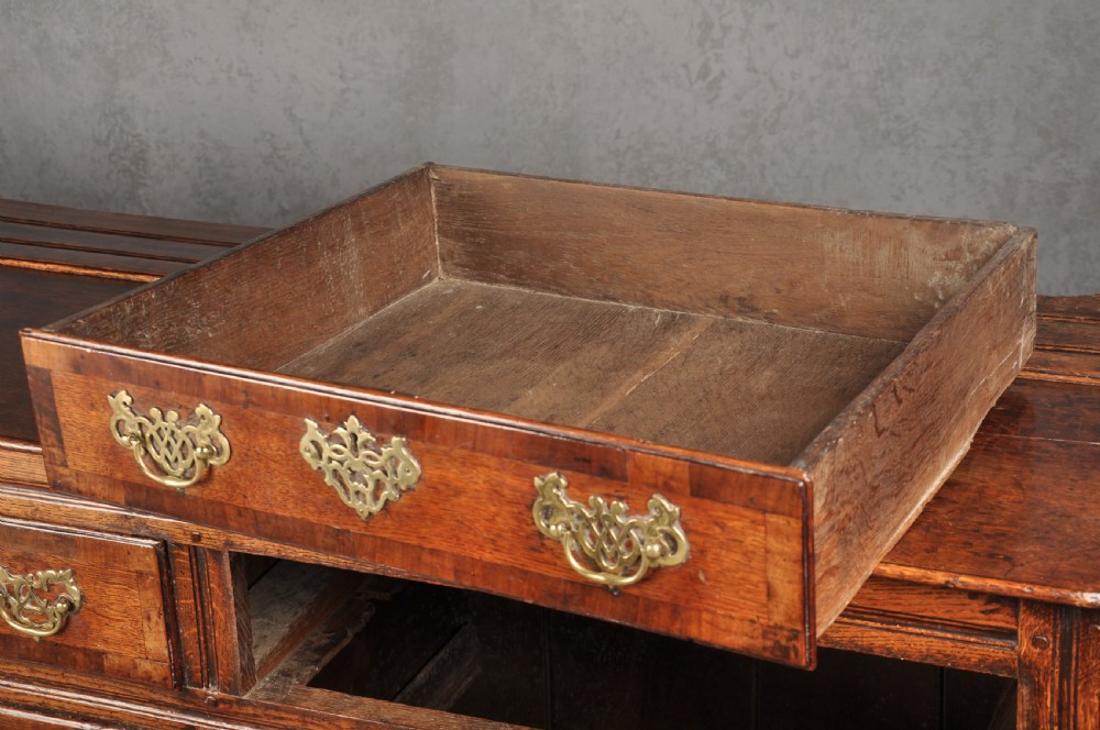 An early 18th-century dark oak dresser base featuring fielded panel cupboard doors, multiple drawers, and brass backplate handles. View XV