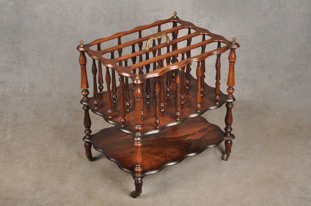 An early nineteenth-century dark rosewood Canterbury music stand featuring vertical spindle dividers, a brass central handle, a lower tier, turned legs, and brass castors.
