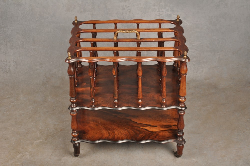 An early nineteenth-century dark rosewood Canterbury music stand featuring vertical spindle dividers, a brass central handle, a lower tier, turned legs, and brass castors. View II