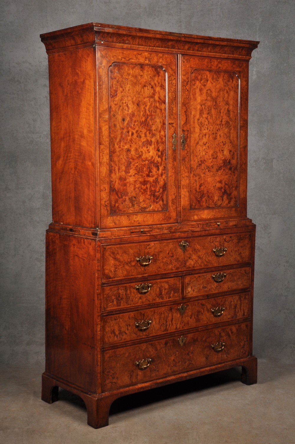An early eighteenth-century burr walnut secretaire cabinet featuring double upper doors and a lower chest of drawers with brass handles.