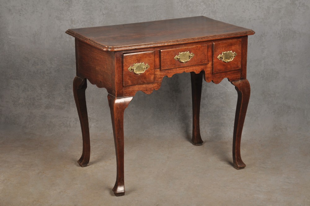 An early 18th-century walnut lowboy side table featuring three drawers with brass handles, a shaped apron, and four cabriole legs.