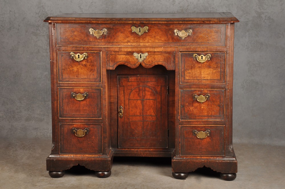 An early 18th-century walnut kneehole desk featuring crossbanded drawer fronts, brass drop handles, a central recessed cupboard, and turned bun feet.