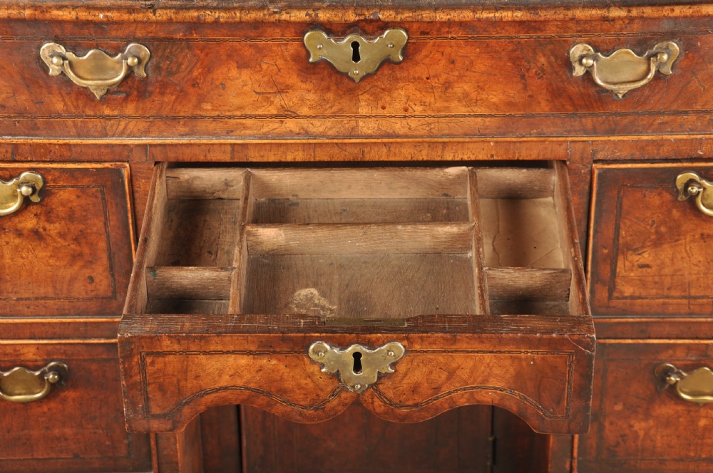 An early 18th-century walnut kneehole desk featuring crossbanded drawer fronts, brass drop handles, a central recessed cupboard, and turned bun feet. View X