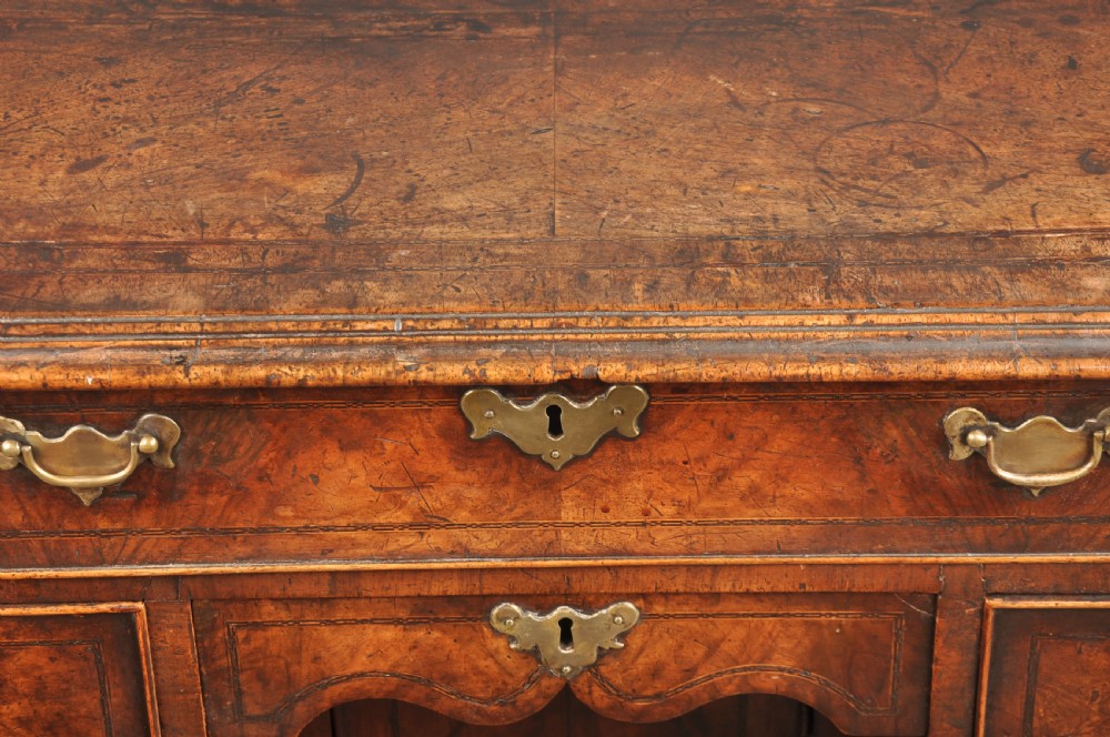 An early 18th-century walnut kneehole desk featuring crossbanded drawer fronts, brass drop handles, a central recessed cupboard, and turned bun feet. View XI