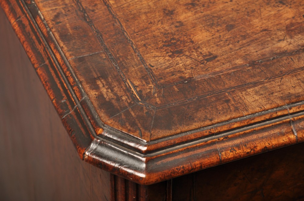 An early 18th-century walnut kneehole desk featuring crossbanded drawer fronts, brass drop handles, a central recessed cupboard, and turned bun feet. View XVI