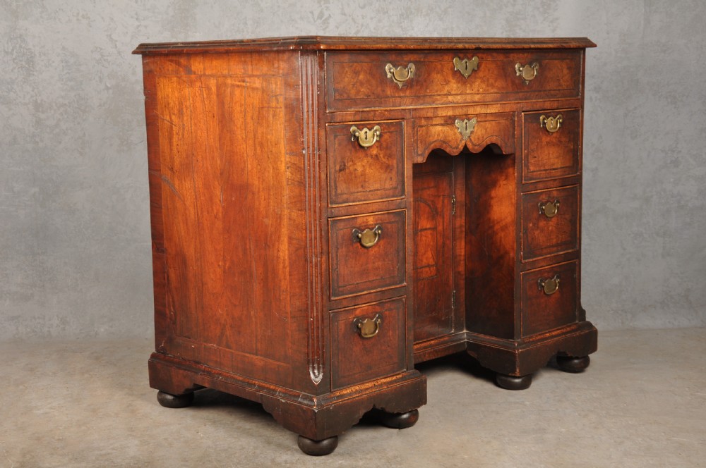 An early 18th-century walnut kneehole desk featuring crossbanded drawer fronts, brass drop handles, a central recessed cupboard, and turned bun feet. View XVII