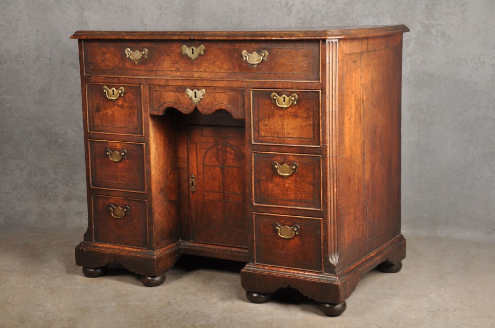 An early 18th-century walnut kneehole desk featuring crossbanded drawer fronts, brass drop handles, a central recessed cupboard, and turned bun feet. View XVIII