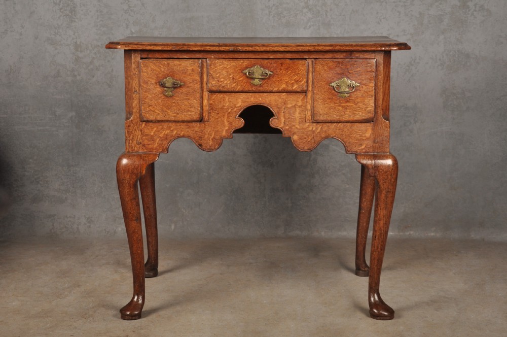 An early eighteenth-century oak lowboy side table featuring three drawers with brass handles, a shaped apron, and four cabriole legs with pad feet.
