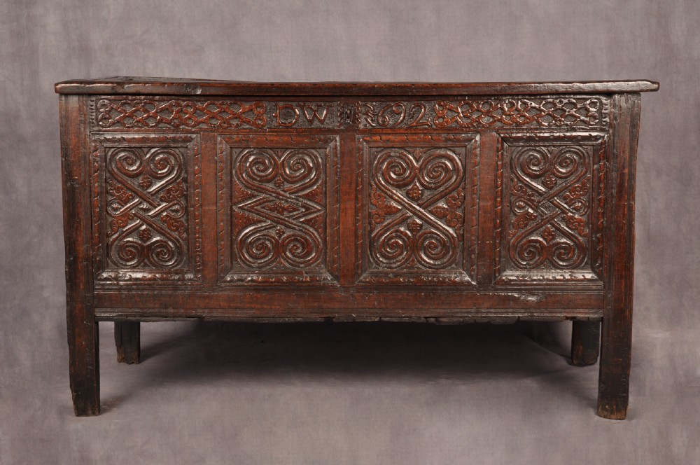 A seventeenth-century dark oak coffer featuring four carved front panels with scroll motifs, a carved top rail dated 1692 with initials DW, and a hinged lid.