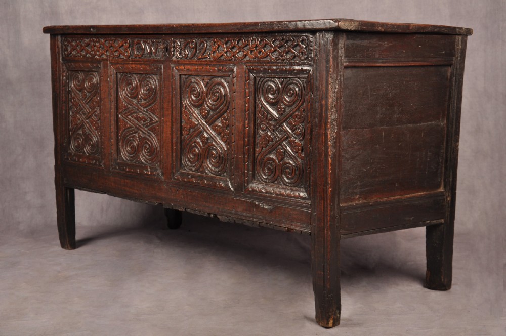 A seventeenth-century dark oak coffer featuring four carved front panels with scroll motifs, a carved top rail dated 1692 with initials DW, and a hinged lid. View IV