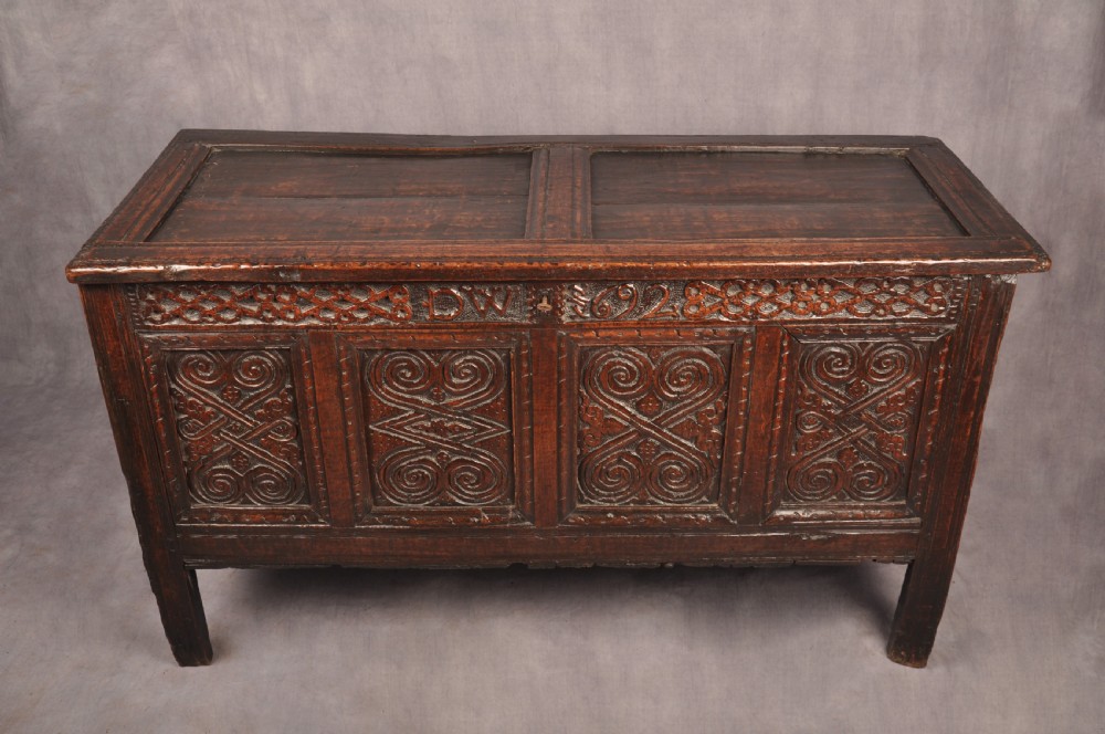 A seventeenth-century dark oak coffer featuring four carved front panels with scroll motifs, a carved top rail dated 1692 with initials DW, and a hinged lid. View V
