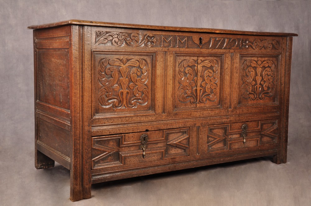 An 18th-century carved oak mule chest featuring three relief-carved front panels, a carved dated top frieze reading 1712, and two lower geometric drawers. View II
