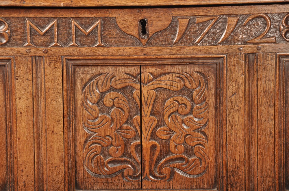 An 18th-century carved oak mule chest featuring three relief-carved front panels, a carved dated top frieze reading 1712, and two lower geometric drawers. View IV