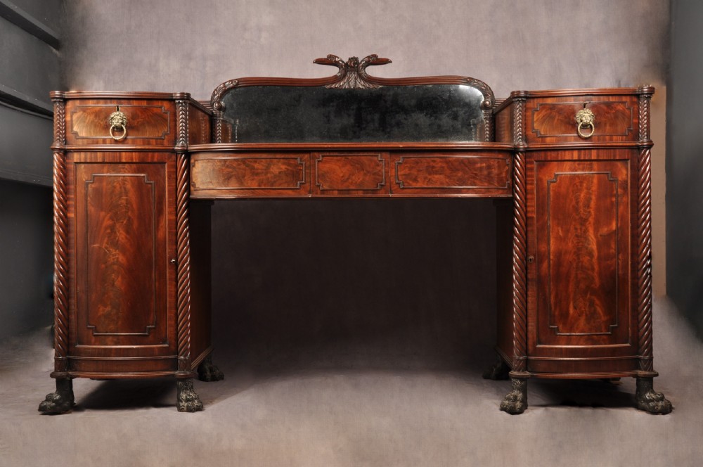 An antique Regency mahogany sideboard featuring flame mahogany panel doors, brass lion mask ring handles, spiral turned columns, hairy paw feet, and a central backboard mirror.