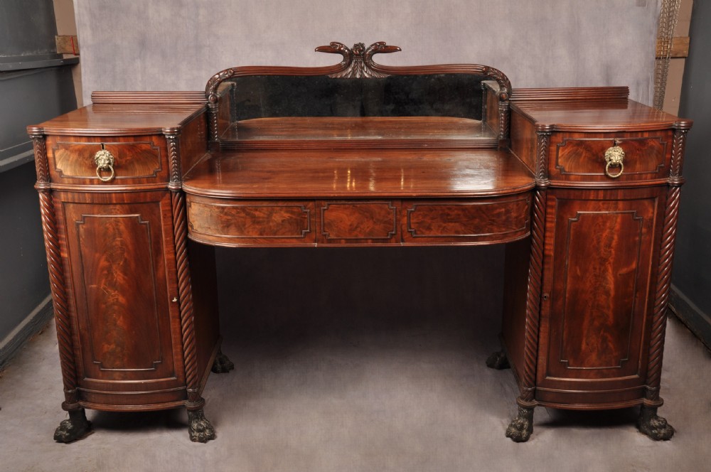 An antique Regency mahogany sideboard featuring flame mahogany panel doors, brass lion mask ring handles, spiral turned columns, hairy paw feet, and a central backboard mirror. View II