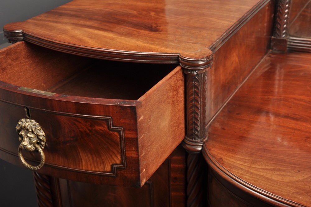 An antique Regency mahogany sideboard featuring flame mahogany panel doors, brass lion mask ring handles, spiral turned columns, hairy paw feet, and a central backboard mirror. View X