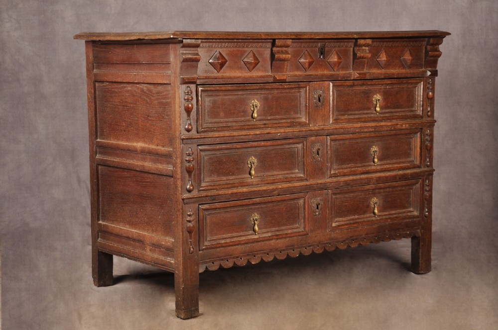 A 17th-century dark oak chest of drawers featuring geometric mouldings, split baluster details, brass drop handles, and a scalloped apron.