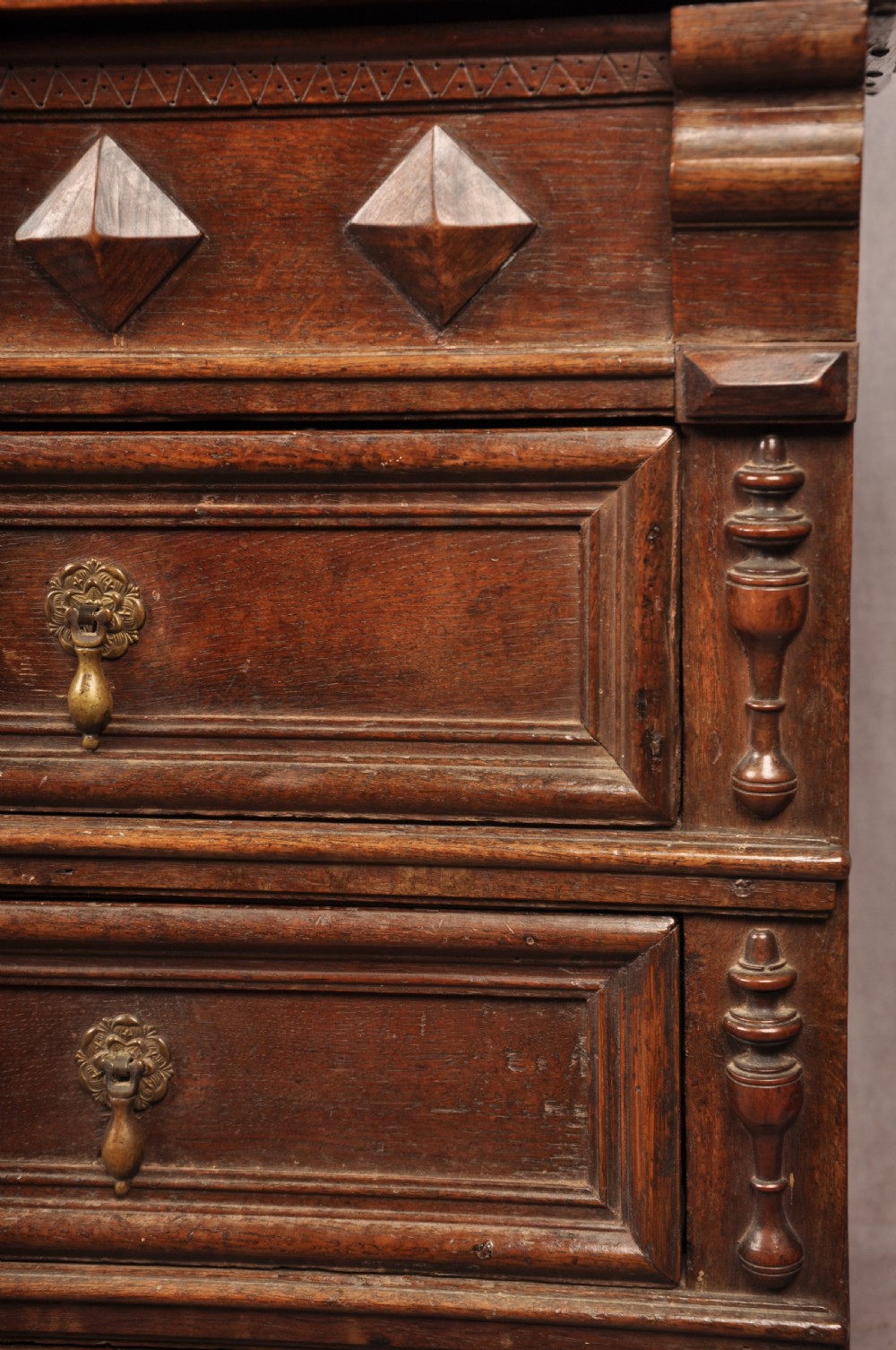 A 17th-century dark oak chest of drawers featuring geometric mouldings, split baluster details, brass drop handles, and a scalloped apron. View VIII