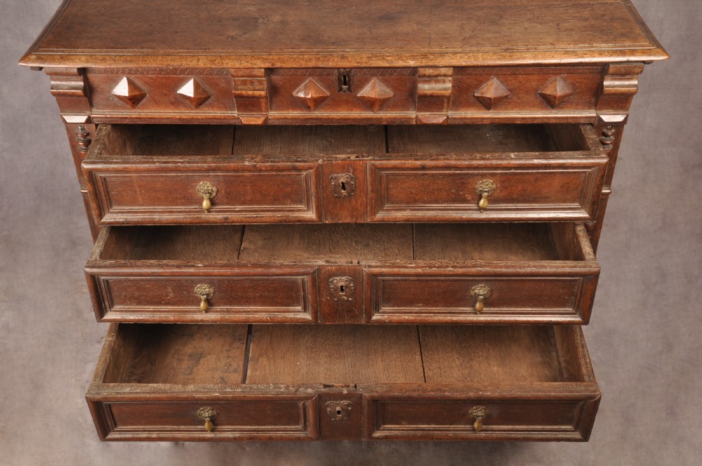A 17th-century dark oak chest of drawers featuring geometric mouldings, split baluster details, brass drop handles, and a scalloped apron. View X