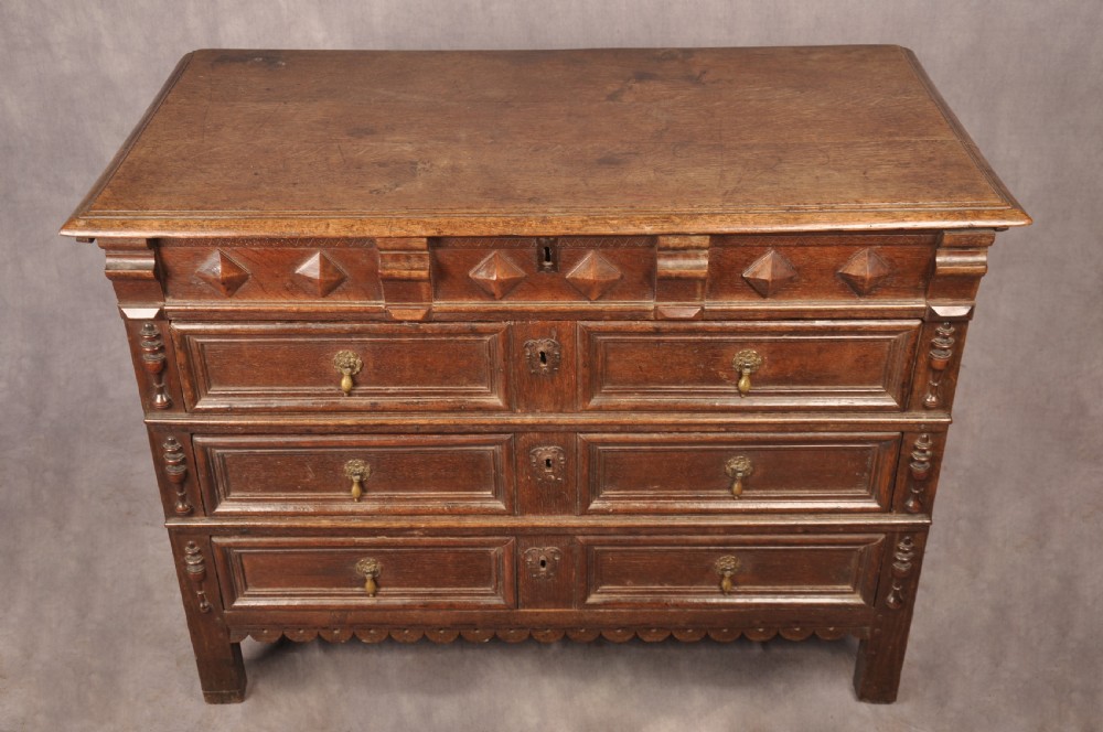 A 17th-century dark oak chest of drawers featuring geometric mouldings, split baluster details, brass drop handles, and a scalloped apron. View XIV