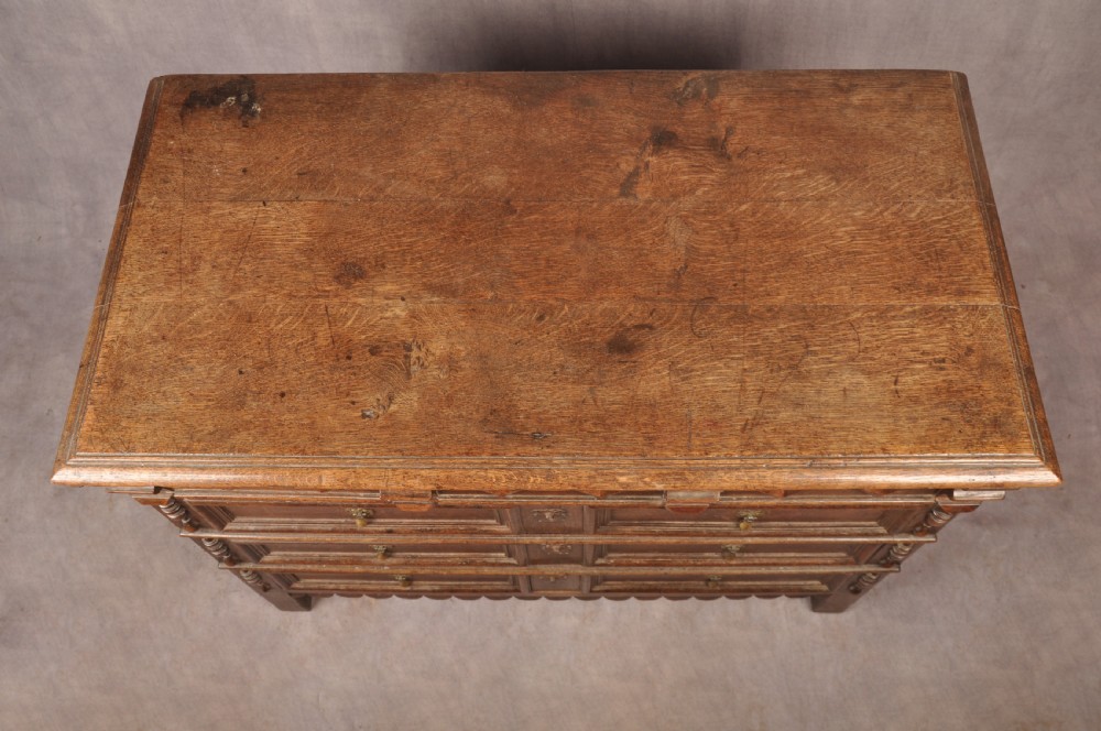 A 17th-century dark oak chest of drawers featuring geometric mouldings, split baluster details, brass drop handles, and a scalloped apron. View XV