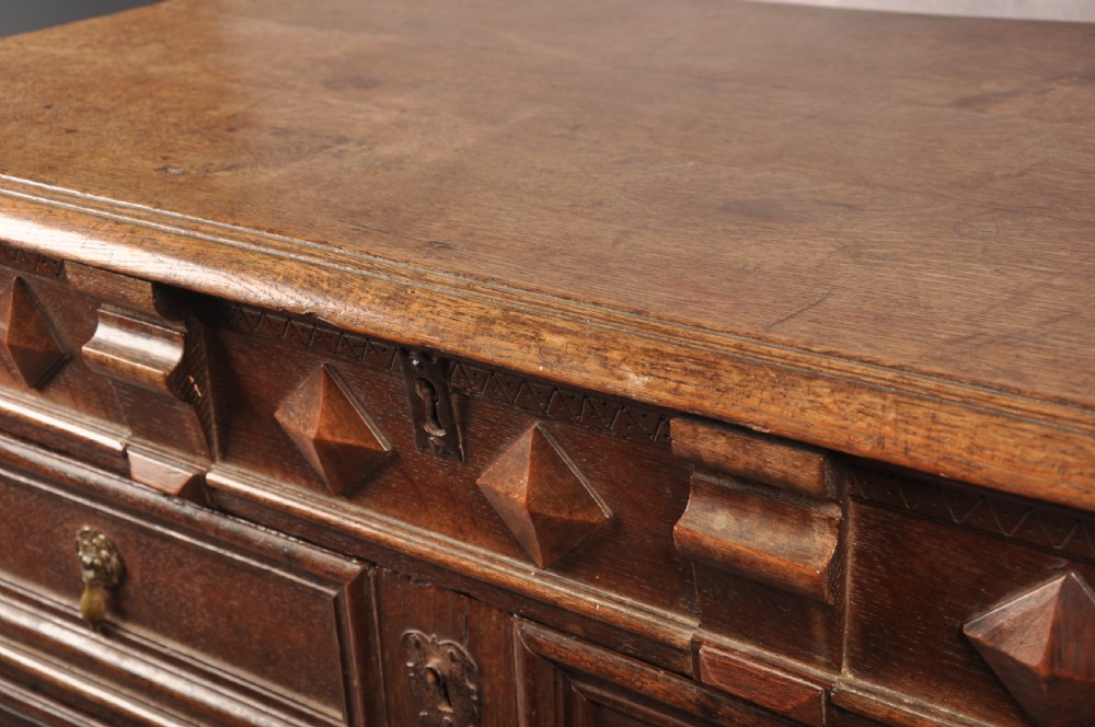 A 17th-century dark oak chest of drawers featuring geometric mouldings, split baluster details, brass drop handles, and a scalloped apron. View XVIII
