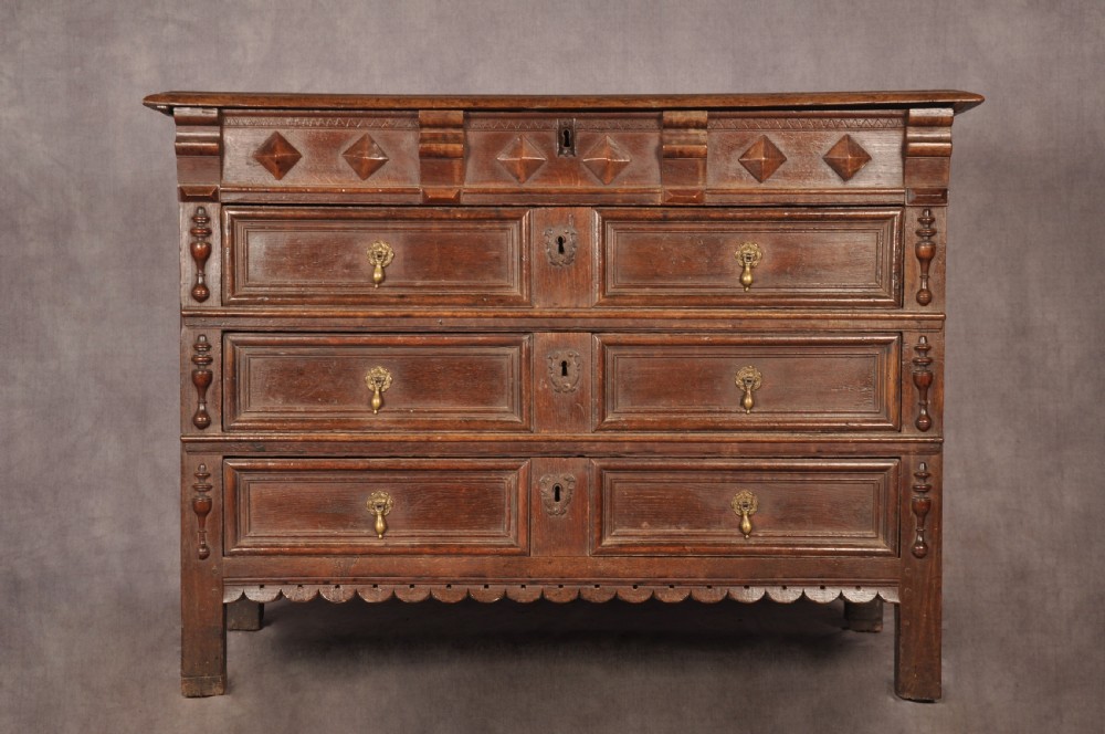 A 17th-century dark oak chest of drawers featuring geometric mouldings, split baluster details, brass drop handles, and a scalloped apron. View XIX