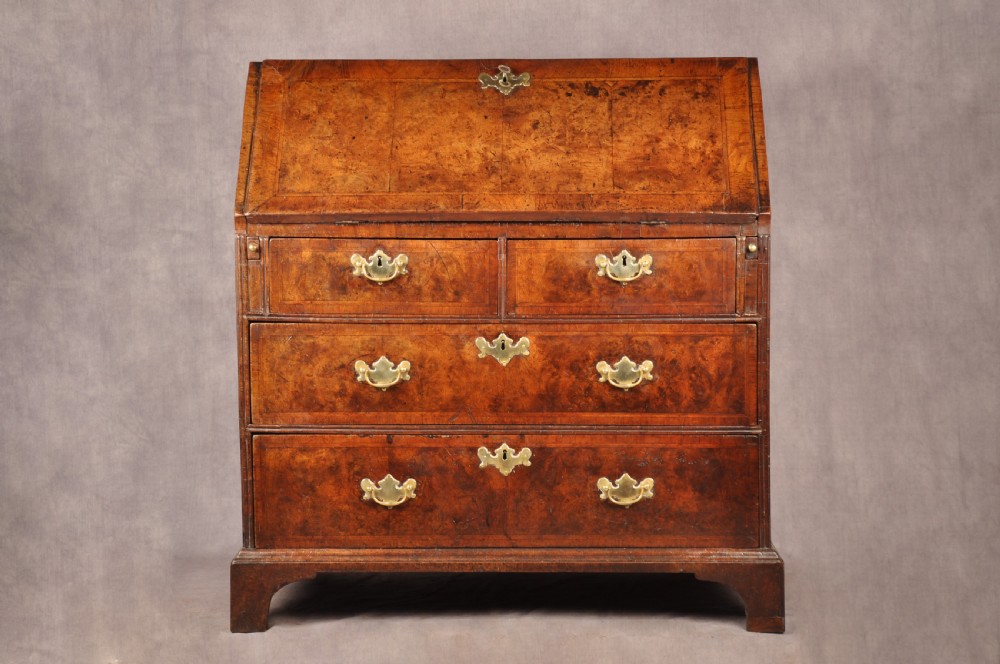 An early 18th-century burr walnut slant-front bureau featuring brass drop handles, keyhole escutcheons, and a chest of drawers base.