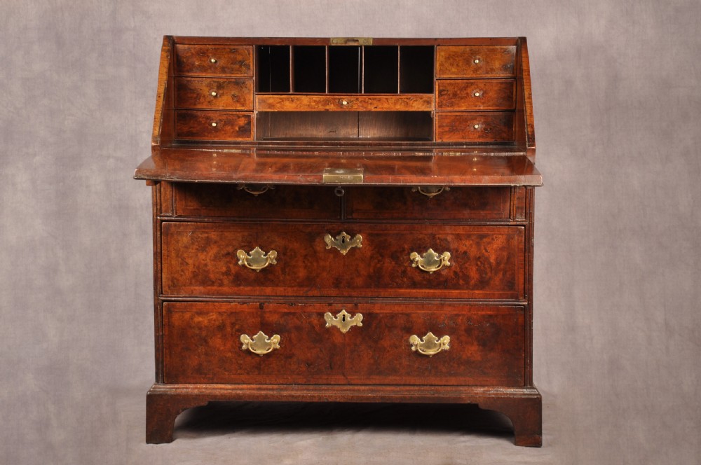 An early 18th-century burr walnut slant-front bureau featuring brass drop handles, keyhole escutcheons, and a chest of drawers base. View V