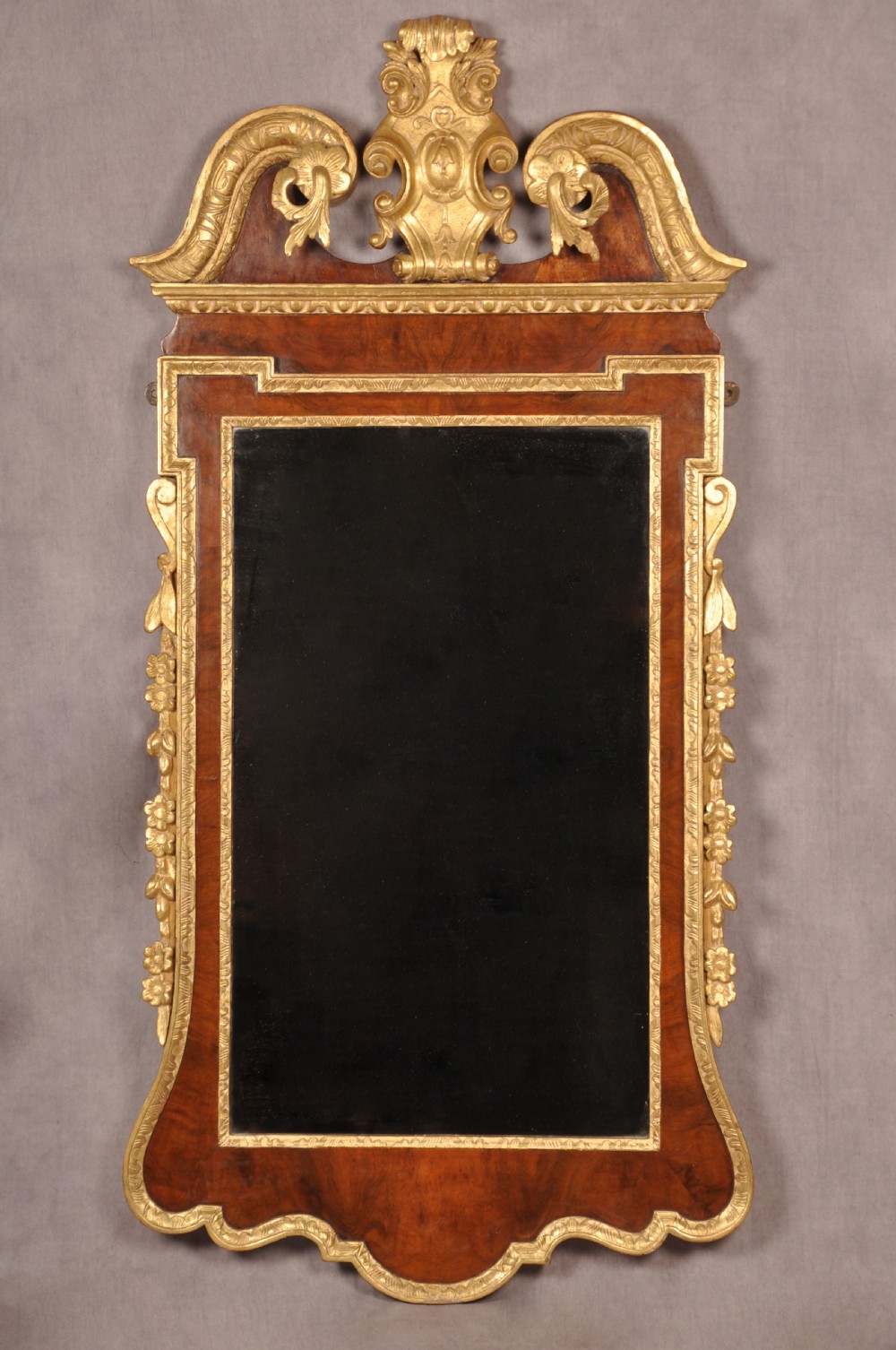 An 18th-century walnut and parcel gilt wall mirror featuring a carved swan-neck pediment, central cartouche crest, and floral side pendants.